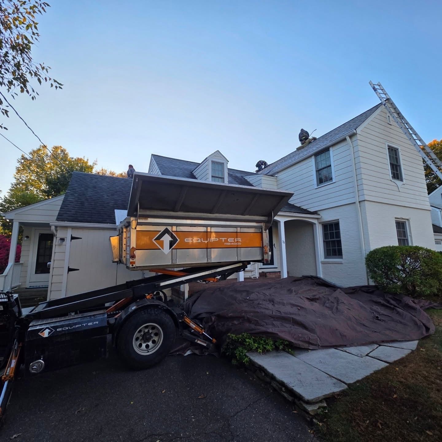 Roofers working on a house; a large dumpster is present in the driveway.