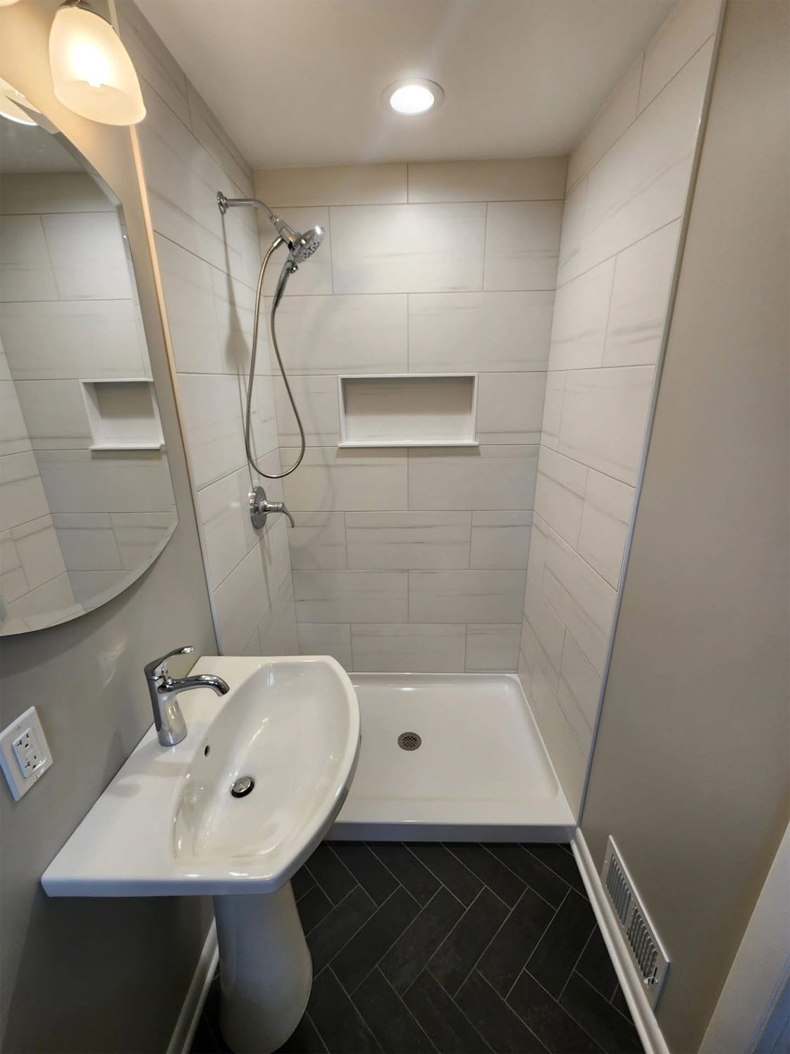 Small bathroom with white tiled shower, pedestal sink, and dark herringbone floor.