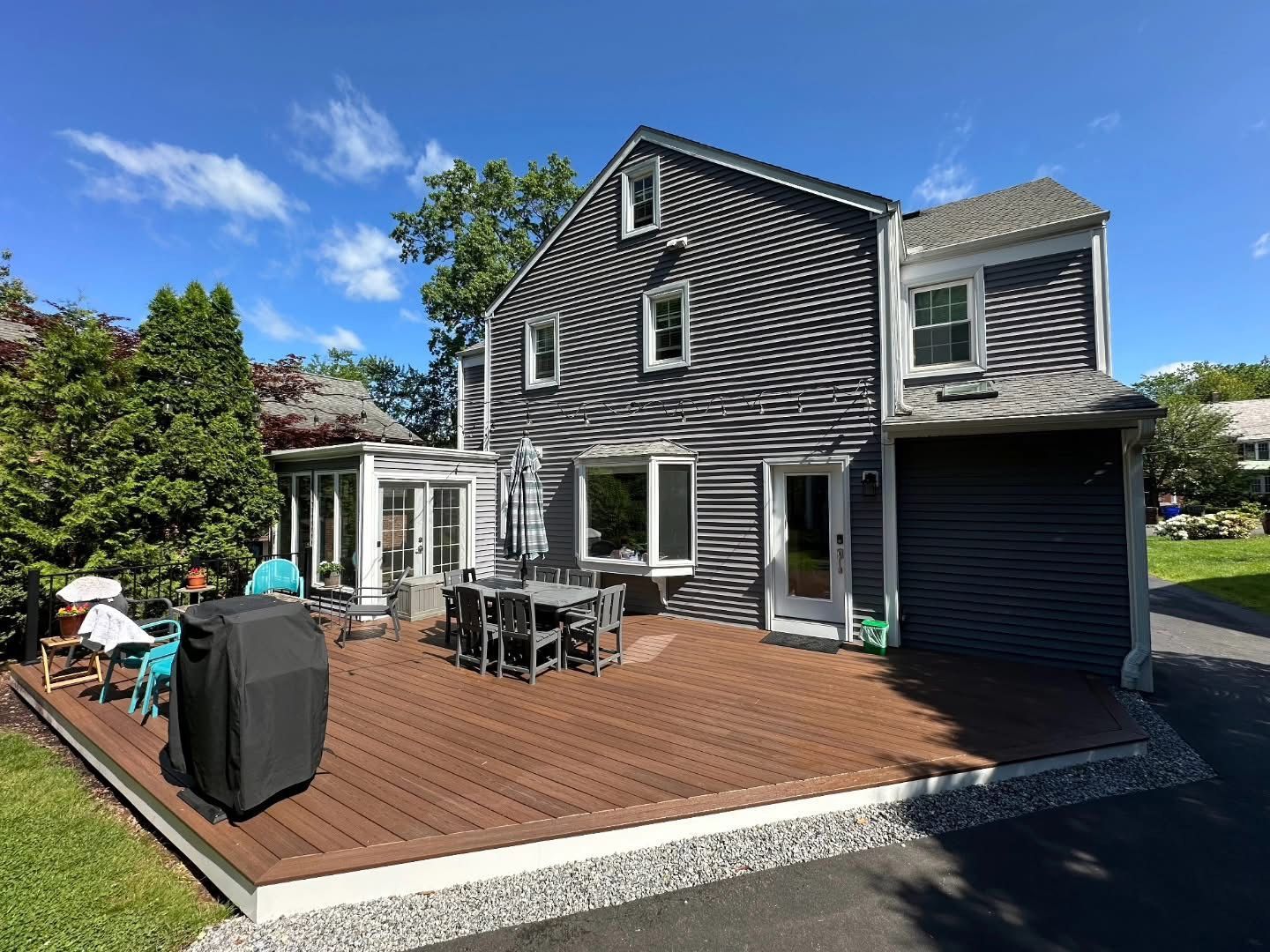 Back view of a two-story gray house with a wooden deck, outdoor table and chairs, and a small attached garage.