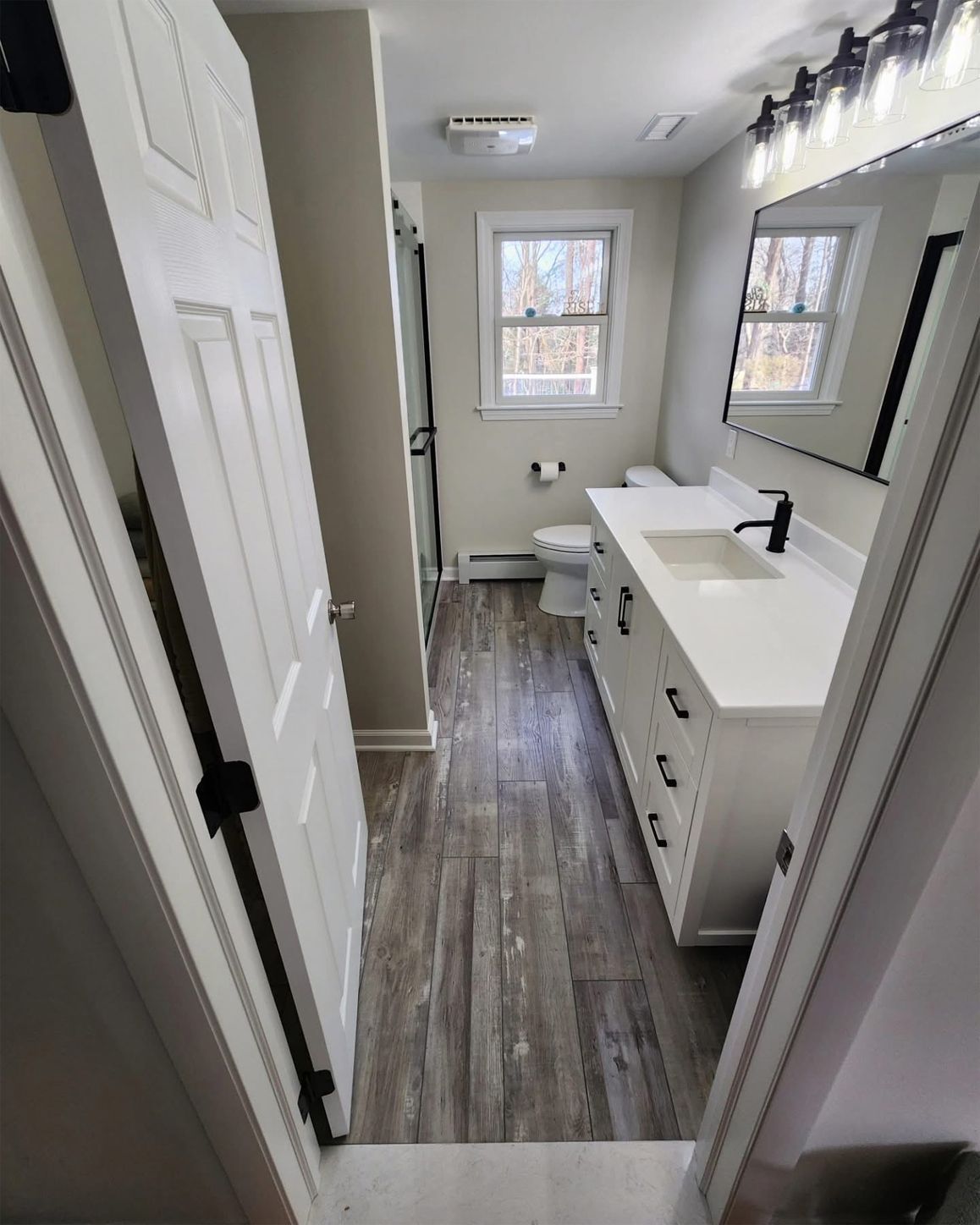 Bathroom with white vanity, black hardware, wood-look flooring, and a window with a view of trees.