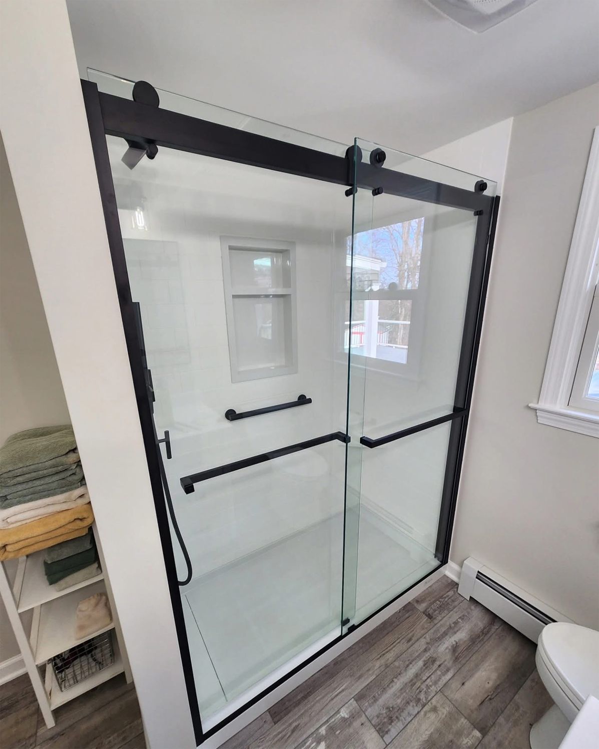 Black-framed sliding glass shower enclosure with grab bars in a white-walled bathroom.