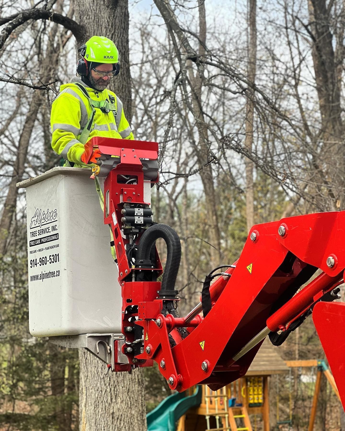 A person in a bright yellow safety vest and helmet operates a red aerial lift bucket amidst a wooded area.