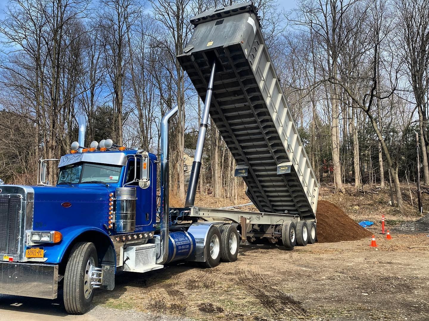 A blue semi-truck with its dump trailer raised to unload a pile of dirt in a wooded area.