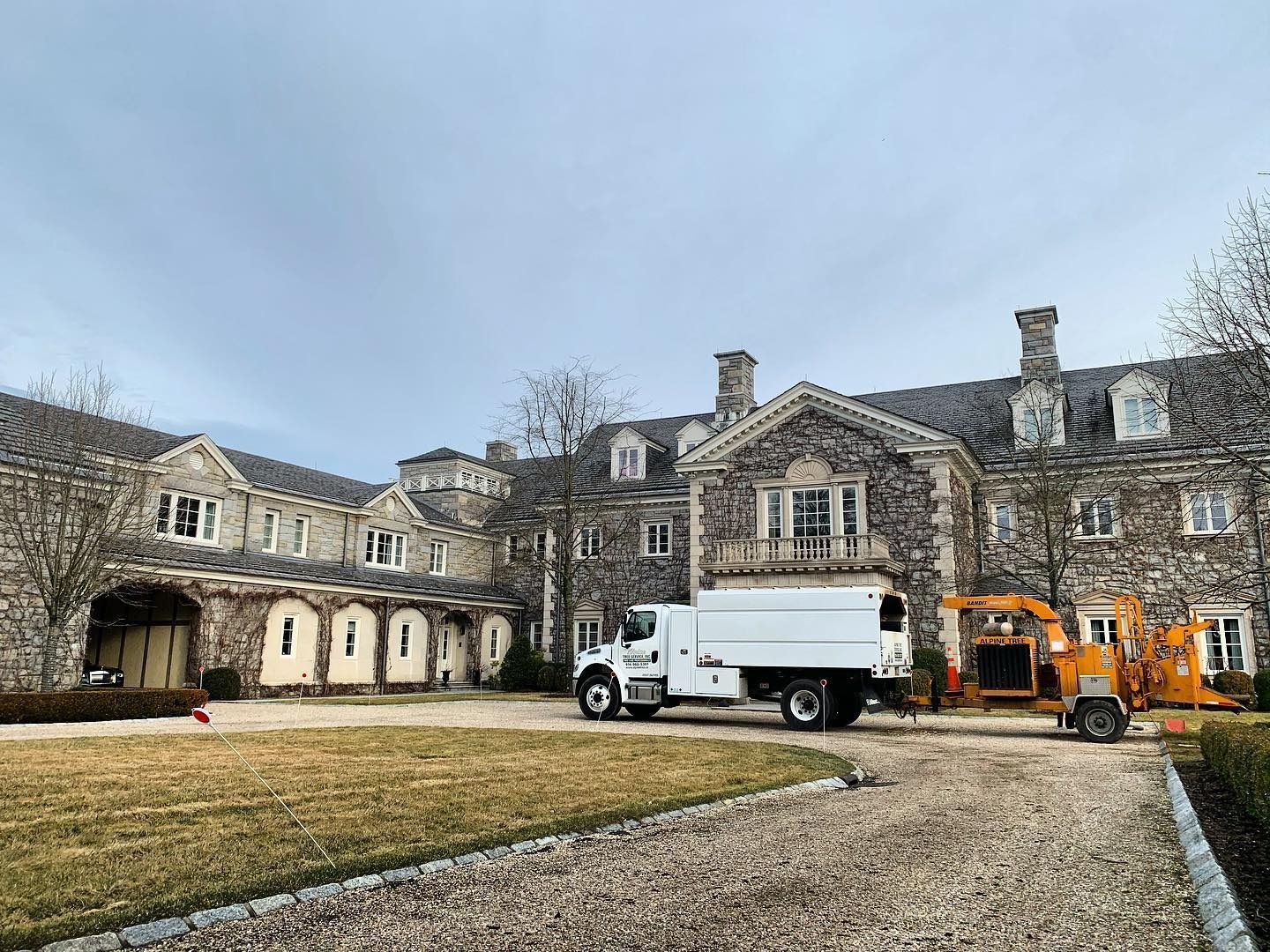 A large stone mansion with a white utility truck and an orange wood chipper parked on the gravel driveway.