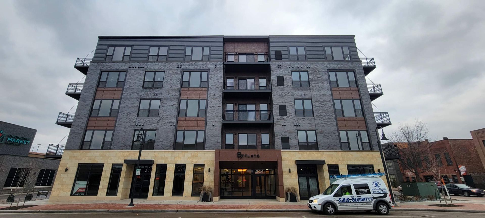 Multi-story apartment building with dark brick facade and street-level commercial space. Cloudy sky above.