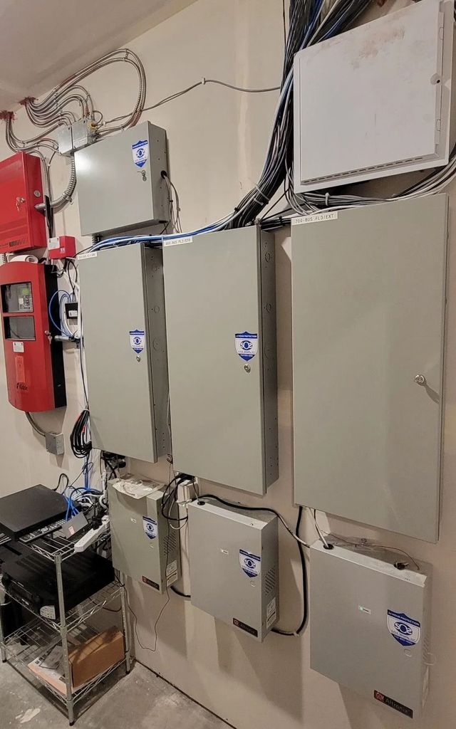 Electrical panel room with multiple gray boxes, wiring, and a red fire alarm system.
