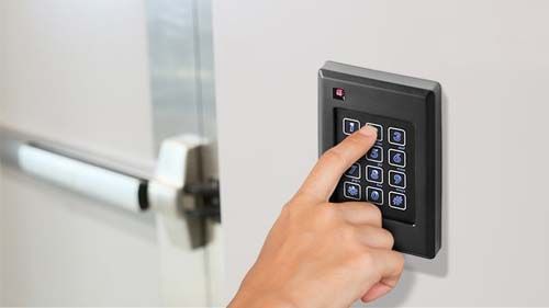 A person's hand pressing keys on a black keypad beside a door with a silver bar handle.