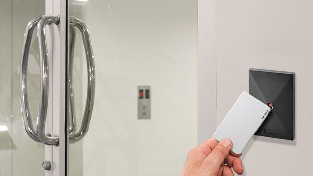 Hand holding card near a black access reader on a white wall; glass door and elevator buttons in background.