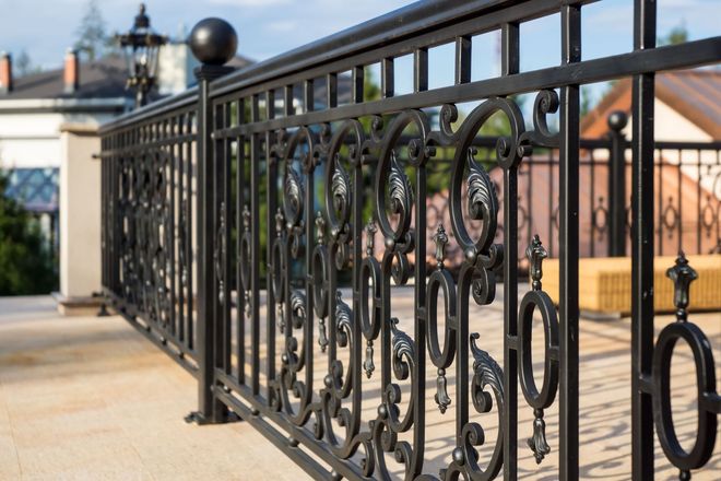A decorative black wrought iron railing borders a paved balcony, with a blurred house and greenery in the background.