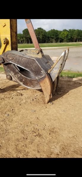 An excavator bucket with a hydraulic thumb attachment resting on a dirt surface outdoors.