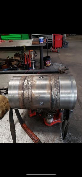 A heavy steel cylinder sits on a shop floor, featuring two welded bands with visible grind marks on the metal surface.