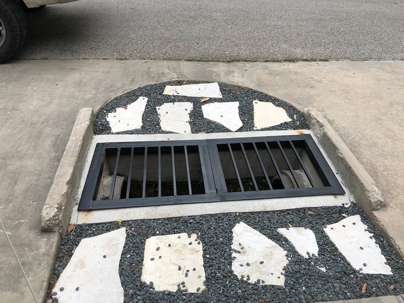 A storm drain with a black metal grate set into a concrete curb, surrounded by gray gravel and decorative stone pavers.