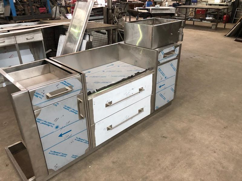 A stainless steel industrial workstation with drawers and cabinets, featuring protective plastic film, in a workshop.