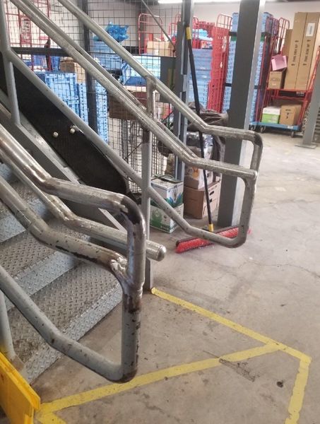 Metal staircase handrails in a warehouse setting, with yellow safety lines painted on the concrete floor.