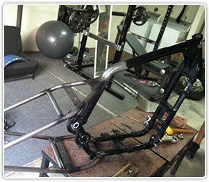 A motorcycle frame sits on a workbench in a home gym, showing exposed metal tubing and black painted sections.