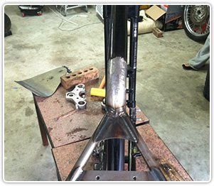 A custom metal gusset welded onto a black motorcycle frame, sitting on a workbench next to a triple tree and tools.