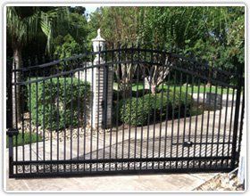 A black metal driveway gate with an arched top, set in a residential landscape with trees and bushes.