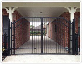 A black metal driveway gate installed between two brick house walls under a covered walkway.