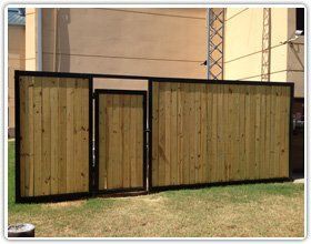 A wooden fence with a centered gate, featuring vertical planks framed by black metal, set against a beige building exterior.