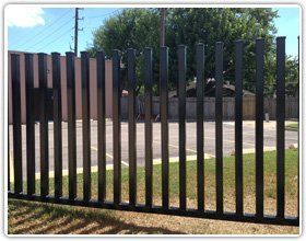 Black metal vertical bar fencing stands in front of a parking lot and a building under a blue sky.