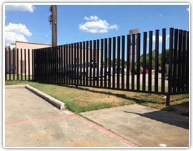 A black steel security fence lines a parking lot under a bright blue sky.