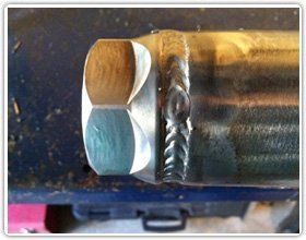 A close-up view of a metal hex nut welded to a steel pipe, showing a clean, stacked-dime TIG weld bead.