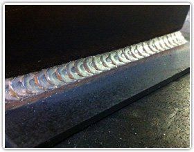 A high-quality, uniform TIG weld bead on dark metal, showing a consistent, stacked-coin pattern along a fillet joint.