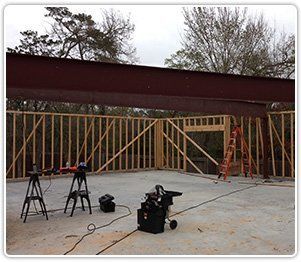 A large steel I-beam is positioned atop wooden wall frames at a construction site with tools on a concrete floor.