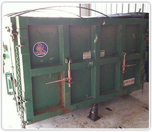 A large, green, rectangular metal industrial waste container or dump trailer with metal latch hardware on its side.