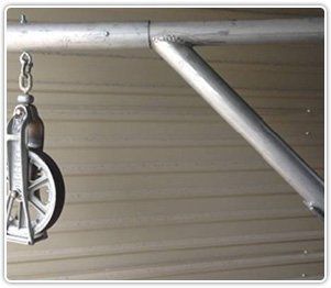 A metal pulley hangs from a horizontal support arm reinforced by a diagonal brace against a ribbed metal wall.
