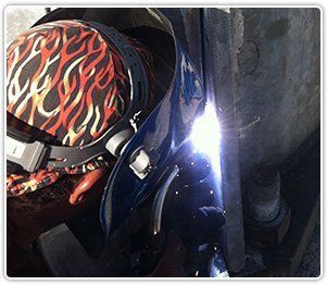 A welder wearing a flame-patterned head covering and protective gear focuses on welding a metal joint, emitting bright light.