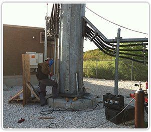 A worker welds the base of a tall metal utility pole in a gravel lot next to a building.