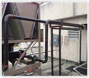 A large industrial chiller unit on a flat roof with black piping connected to a support frame near a building wall.