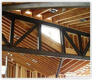 A dark metal truss spans across the interior of a house under construction, with wooden rafters visible in the background.