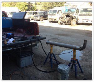 A metal pipe is supported by two yellow stands outdoors, with welding equipment in the bed of a nearby pickup truck.