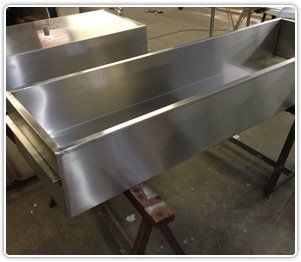 A long, rectangular, polished stainless steel trough resting on wooden sawhorses in a workshop.