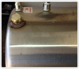 Close-up of a stainless steel water heater tank showing a brass plug and two white pipe fittings on the top surface.