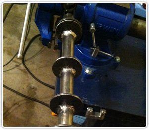 A metal shaft with three welded circular flanges clamped in a blue bench vise.