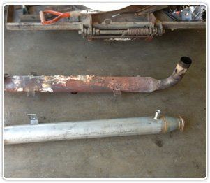 A rusted, older exhaust pipe lies on the ground next to a new, clean, silver replacement pipe.