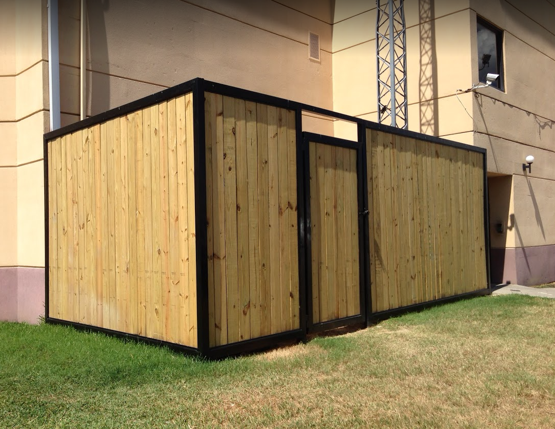 A rectangular outdoor enclosure with vertical wooden planks and a black metal frame stands against a building wall.