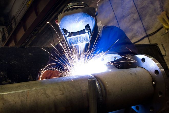 A welder wearing a protective mask joins a metal pipe to a flange as bright sparks fly in a workshop setting.