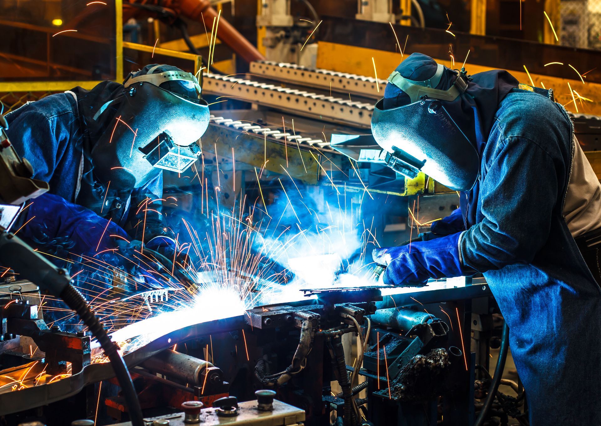 Two workers in protective gear and helmets welding metal, creating bright sparks in an industrial workshop setting.