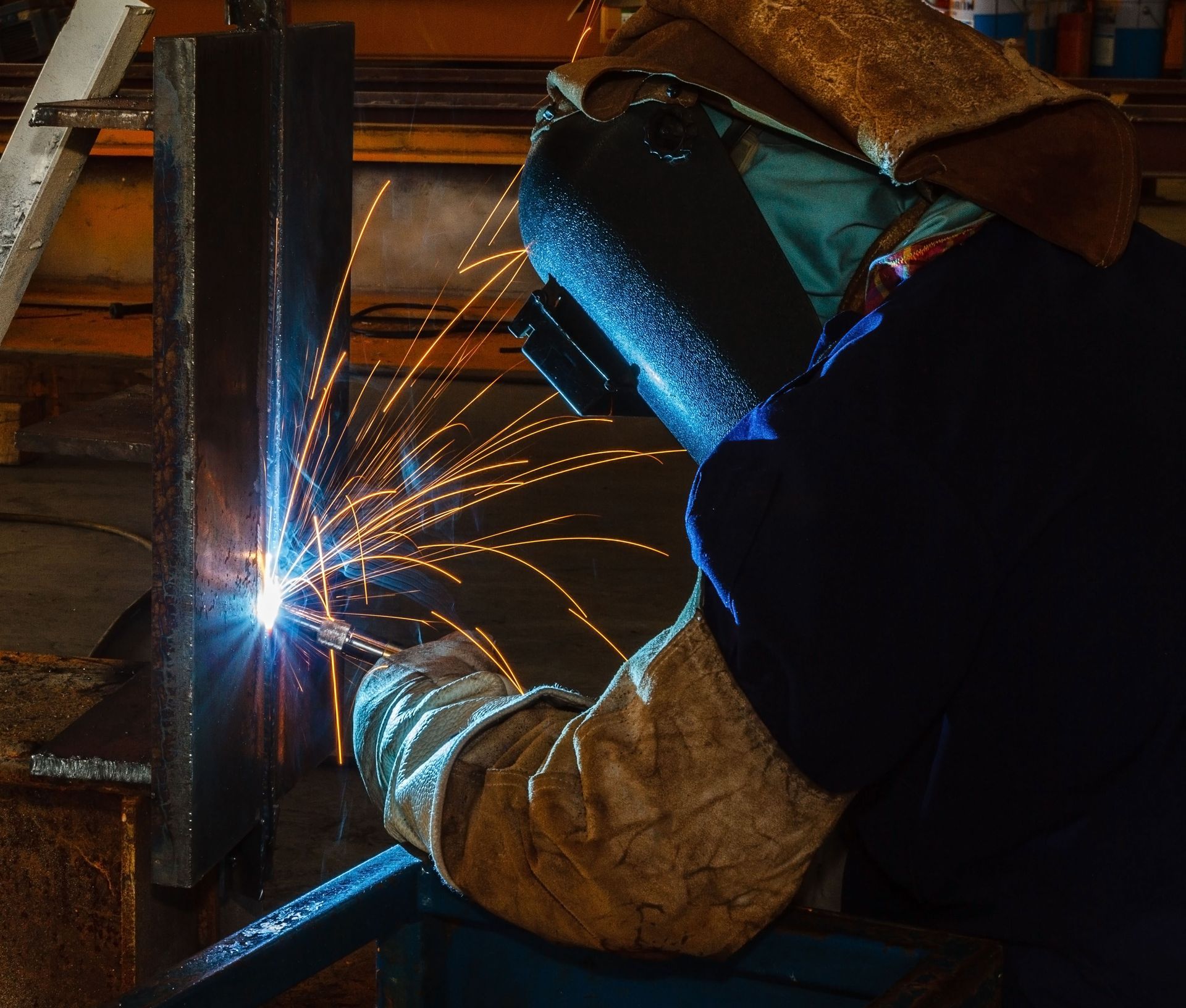 A welder in protective gear uses a torch to join metal pieces, creating bright orange sparks against a dark background.