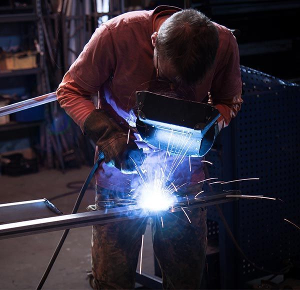 A person wearing a welding helmet and gloves performs metal welding, creating bright sparks in a workshop.