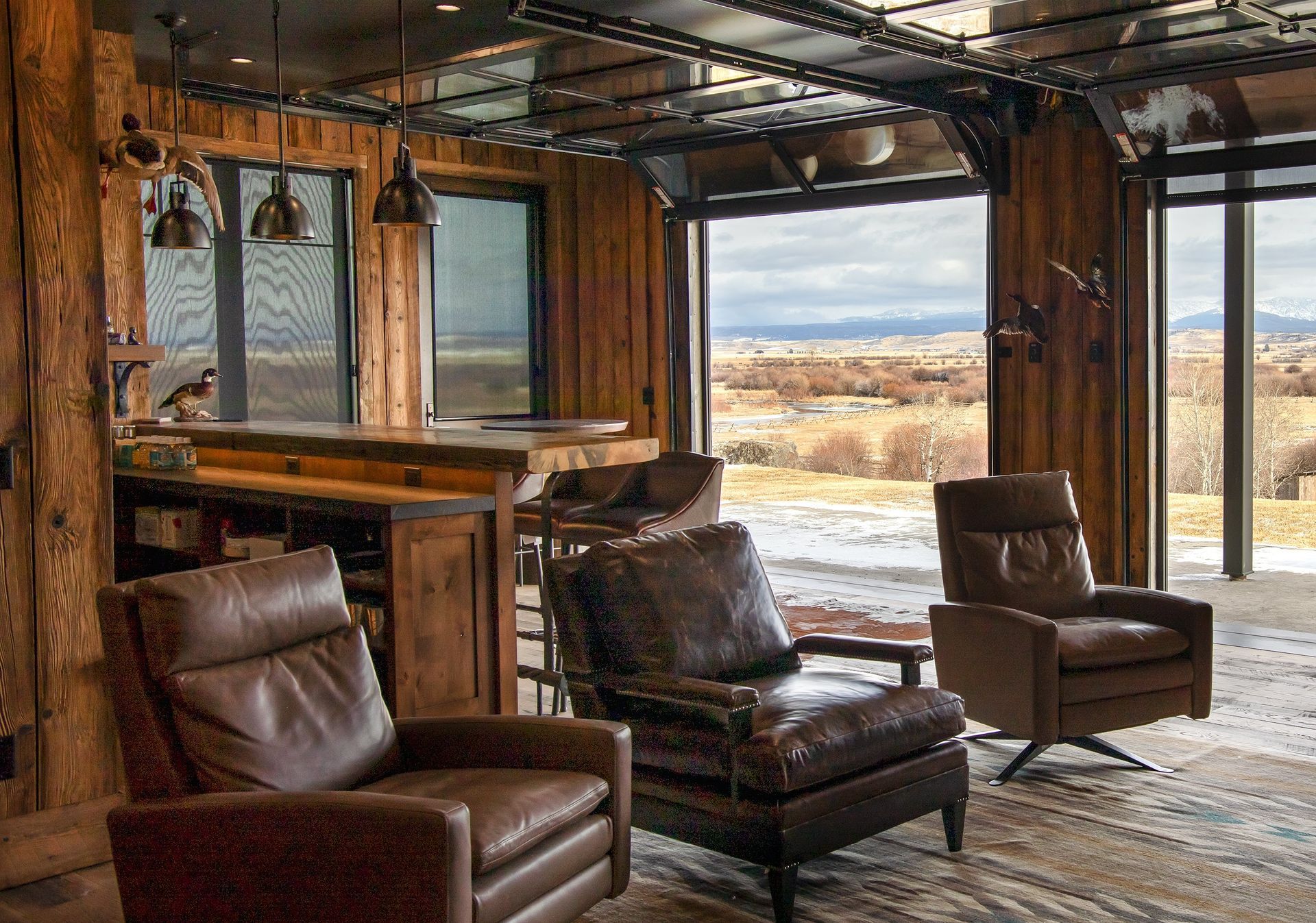 Cozy rustic room with leather chairs, bar, and open garage door to a snowy landscape.