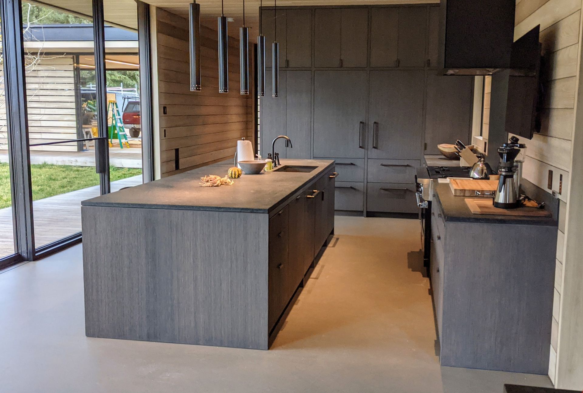 Modern gray kitchen with island, built-in appliances, and sleek hanging lights; large windows and open-air view.