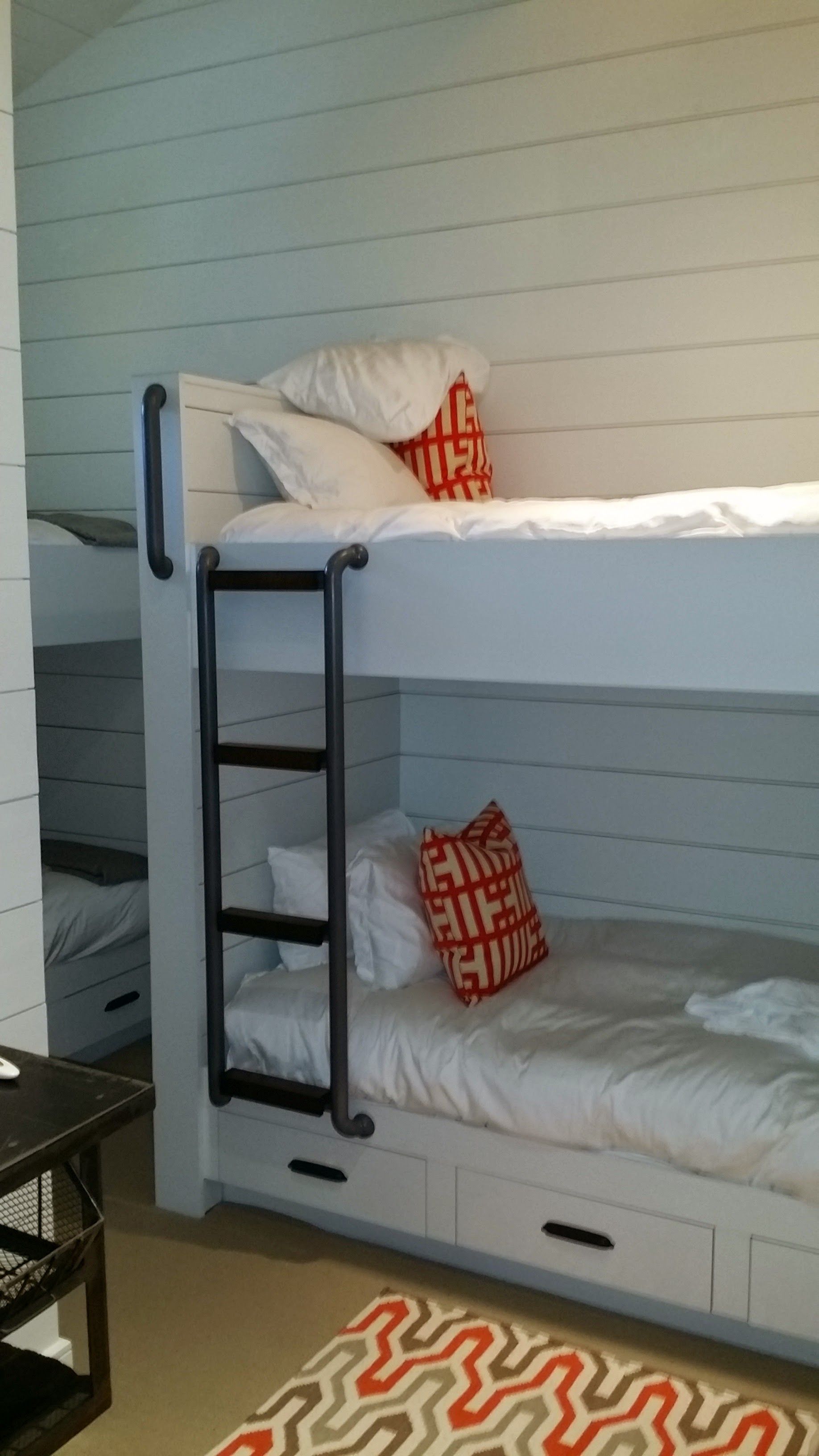 White bunk beds with black ladder and two orange patterned pillows in a room with white horizontal plank walls.