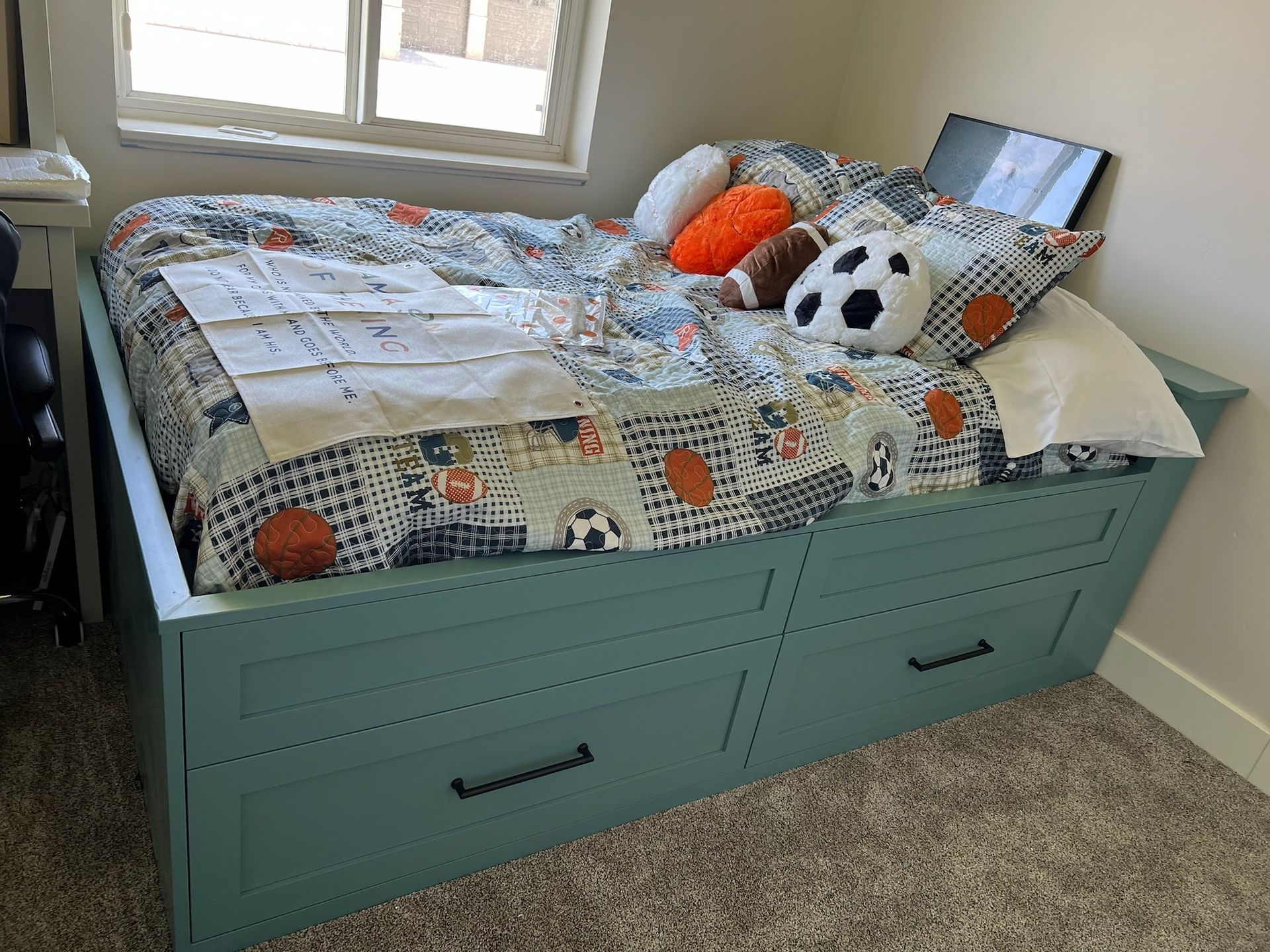 Teal bed with large drawers, sports-themed bedding and pillows, near a window.