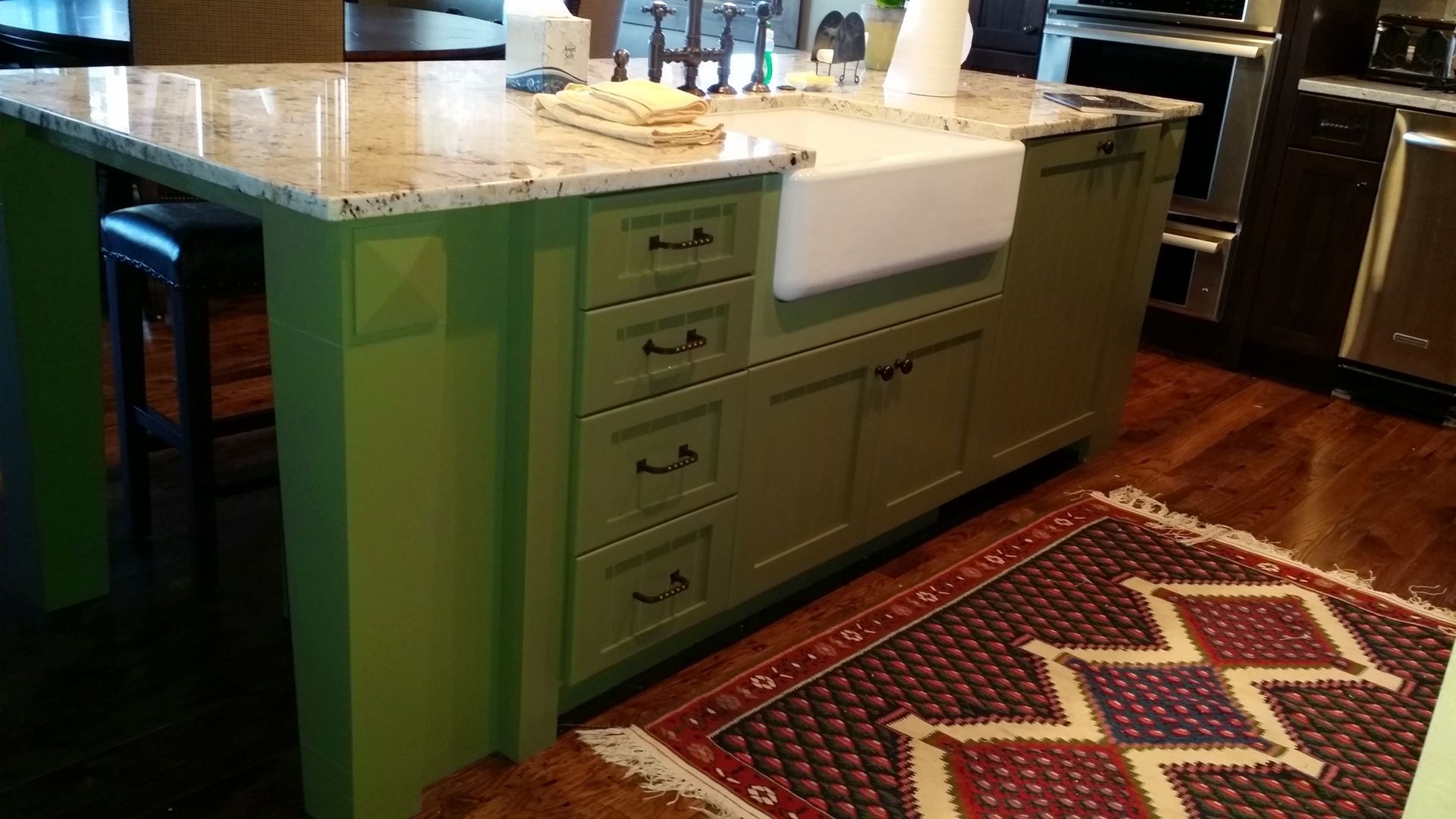 Green kitchen island with drawers and sink, granite countertop, black bar stool, rug.