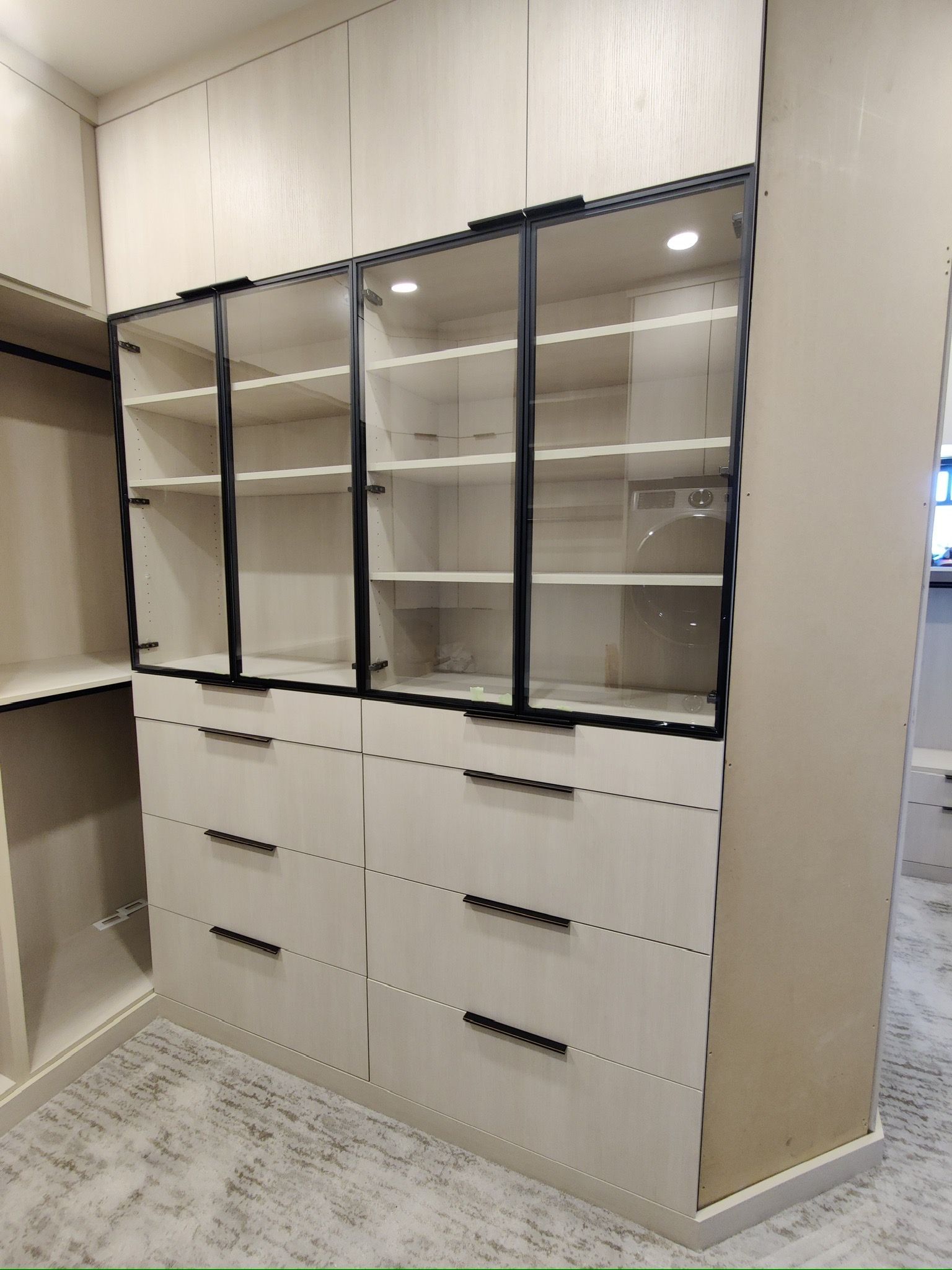Light-colored custom closet with drawers, glass-front shelves, and black trim; carpeted floor.